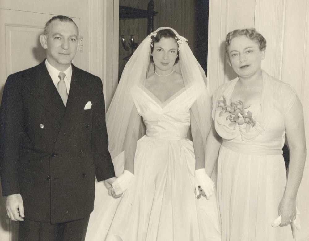 Bertha Rose in her wedding dress and veil, holding hands with her mother and father on either side of her in a doorway.