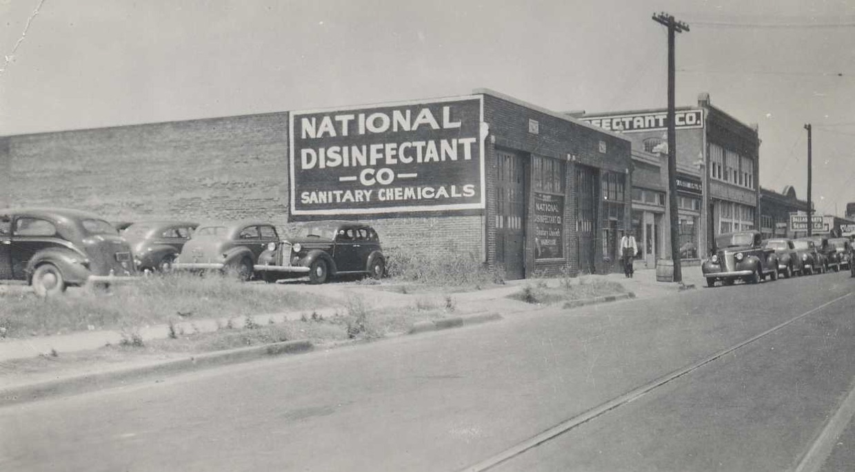 A long brick building with NATIONAL DISINFECTANT CO — SANITARY CHEMICALS painted across its end wall, cars of the period parked along the kerb and a man walking past the loading doors.