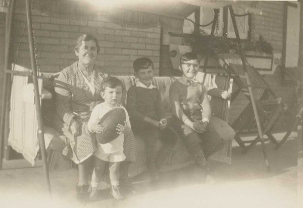 Three young brothers sitting on a porch swing beside their grandmother, the smallest holding a cap on his lap.