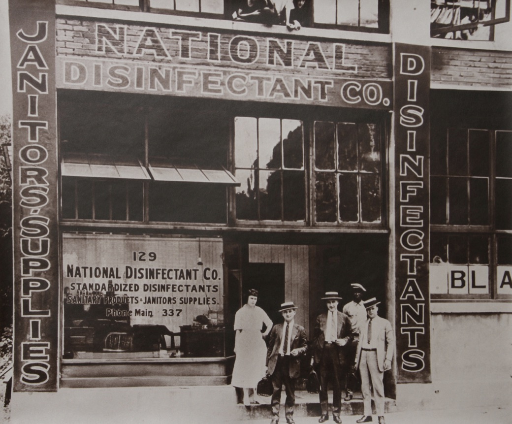 The National Disinfectant Company storefront at 129, photographed around 1918, with four members of staff standing on the step beneath signs reading Janitors&rsquo; Supplies and Disinfectants.